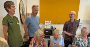 a group of three people stand behind three others who are sat at a table. All are smiling.