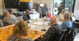 a group of people sit around a table as a man stands one end talking to them.