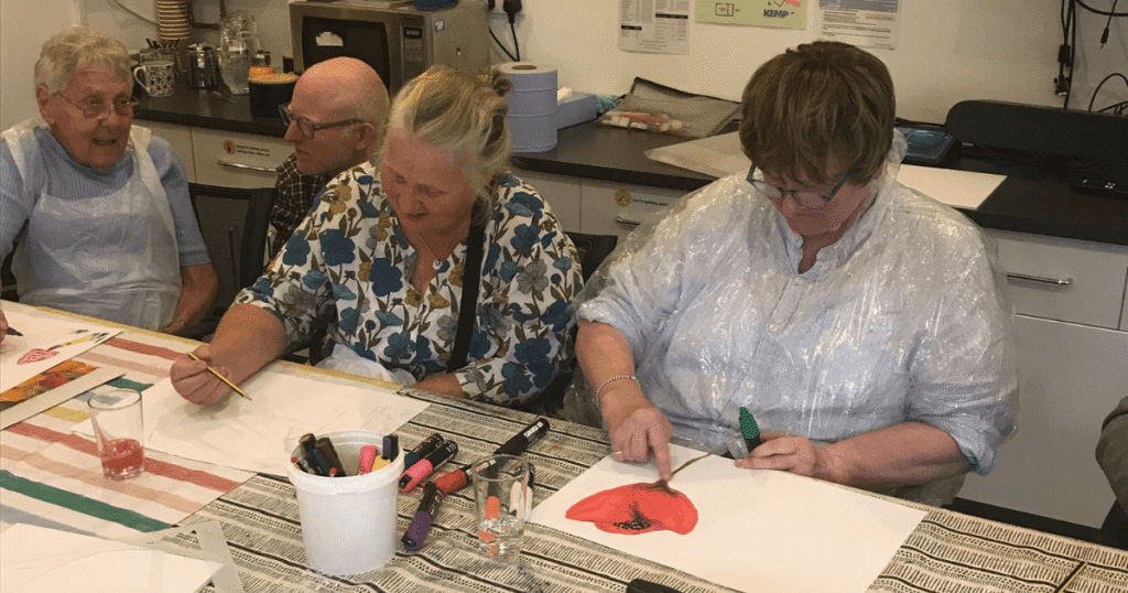 two people sit at a table drawing art such as a poppy and another person is knelt down talking to another lady