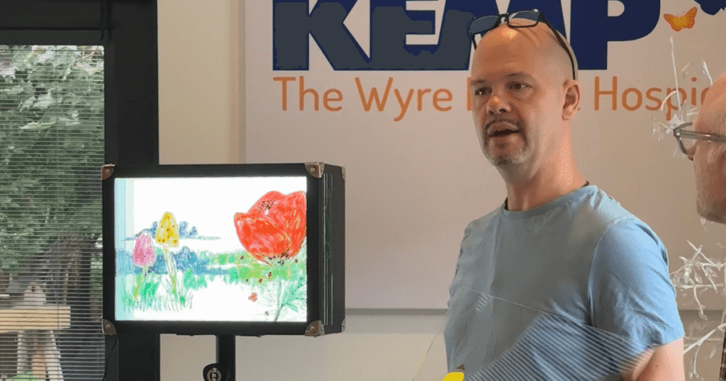 a man in a blue t shirt stands next to a light box with a hand drawn picture of a poppy, mushrooms and green space.