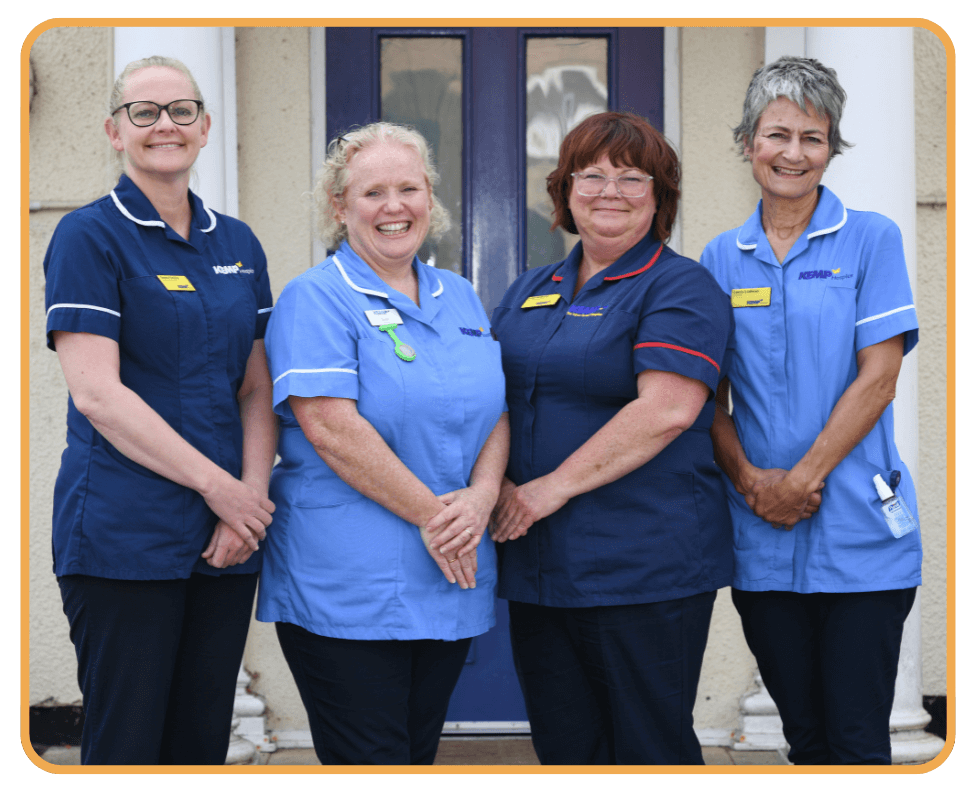 four nurses wearing scrubs stand together in front of a cream colored building with a blue door