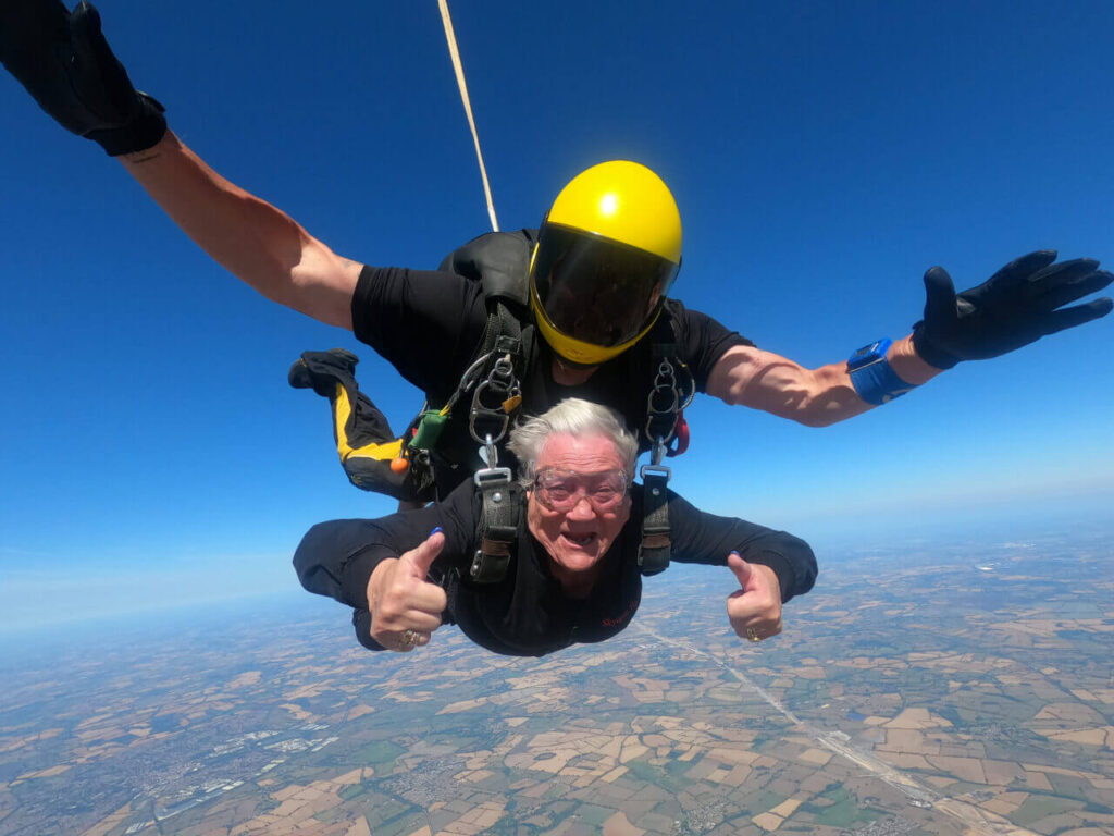 a lady on a tandem skydive falling through he air with her thumbs up at the camera. The background is blue sky and the earth can be seen in the bottom of the picture.