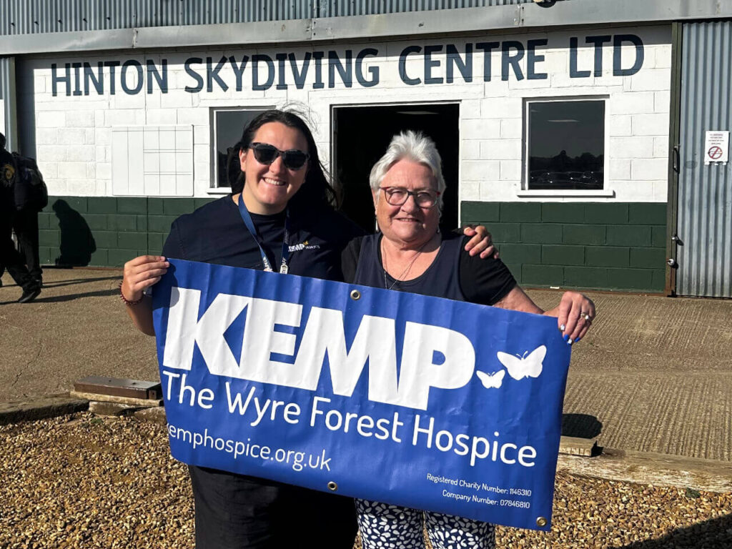 two ladies stand arm in arm holding a kemp hospice blue banner with white logo and text.