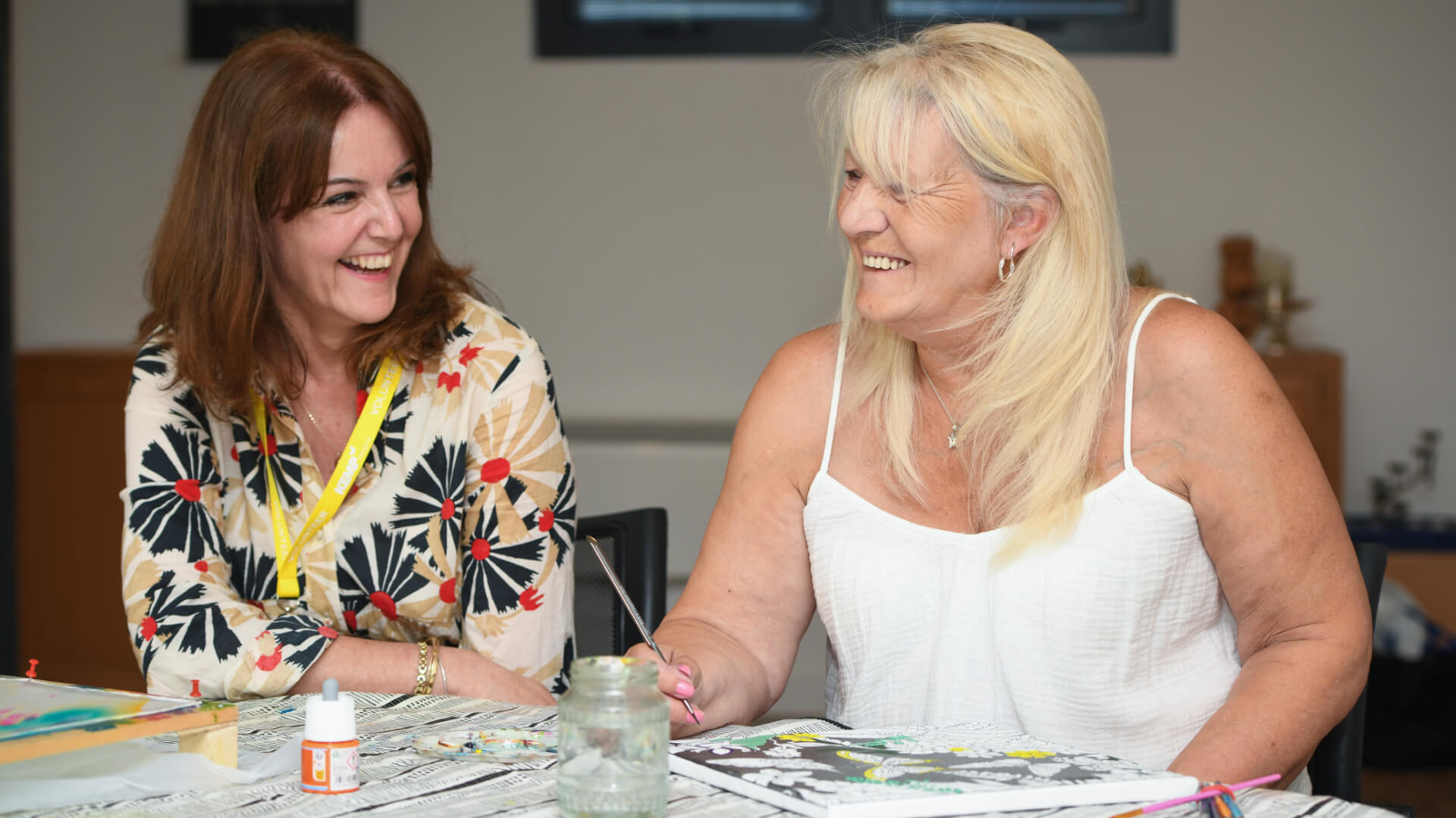 two ladies sit at a table smiling. One is holding a paint brush by a canvas she is painting on.