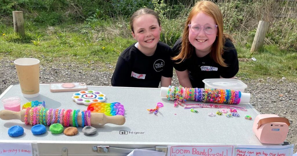 two girls stand behind a table filled with their hand made jewelry for sale