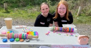two girls stand behind a table filled with their hand made jewelry for sale