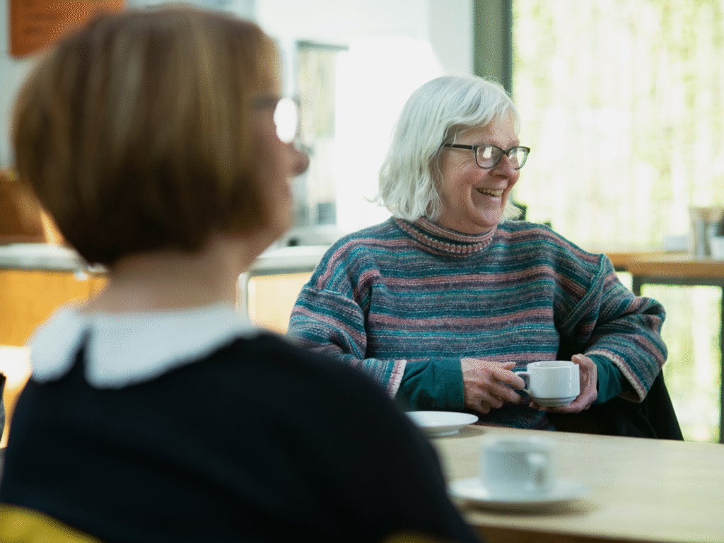 a lady in a striped jumper sits at a table with a coffee cup and saucer smiling whilst enjoying a cup of coffee.