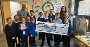 a group of primary school children stand holding a large check with a lady and gentleman at the back