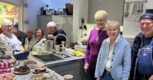 a group of ladies stand next to a kitchen bench with cakes on display for a cake sale.