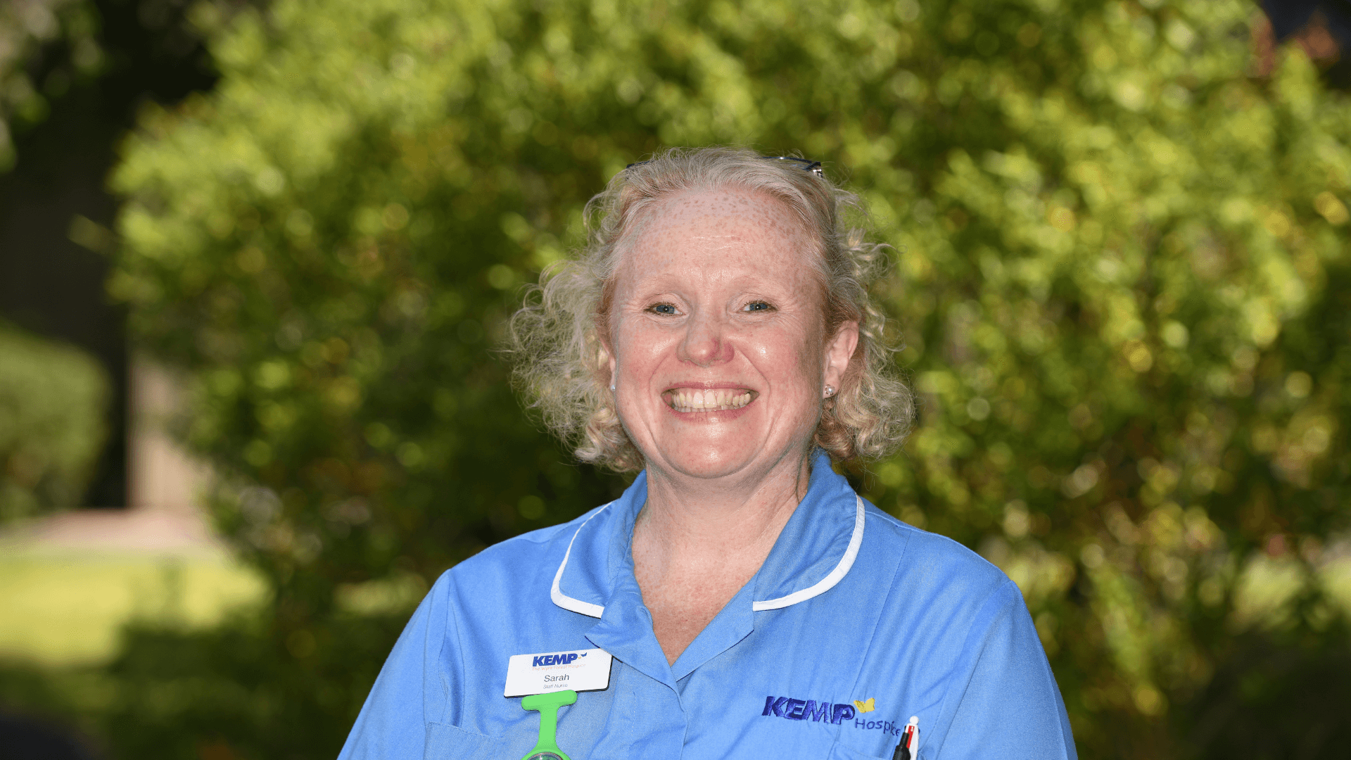 Nurse in kemp hospice uniform with garden bushes in the background