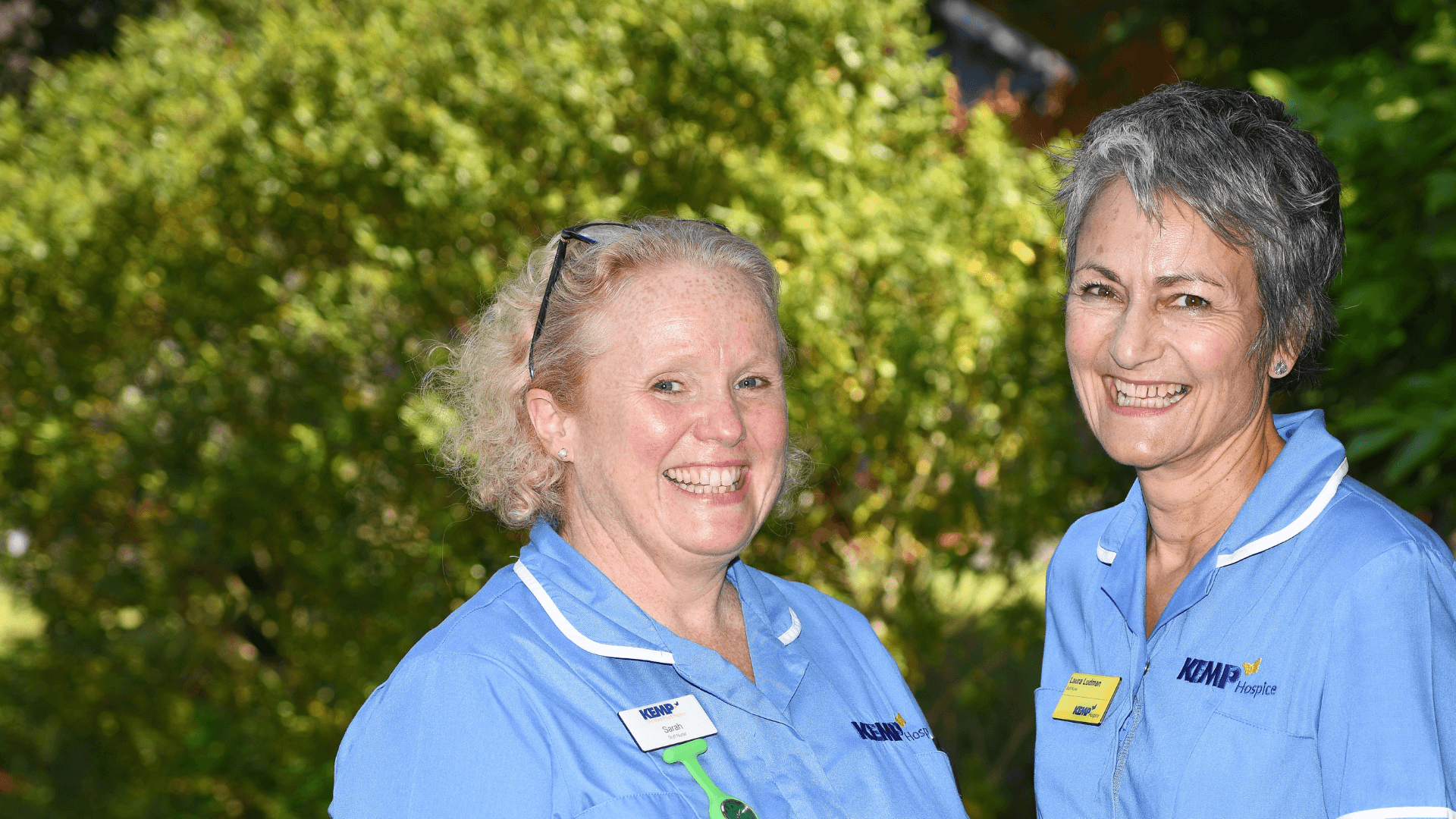 two nurses stand together with a garden area in the background.