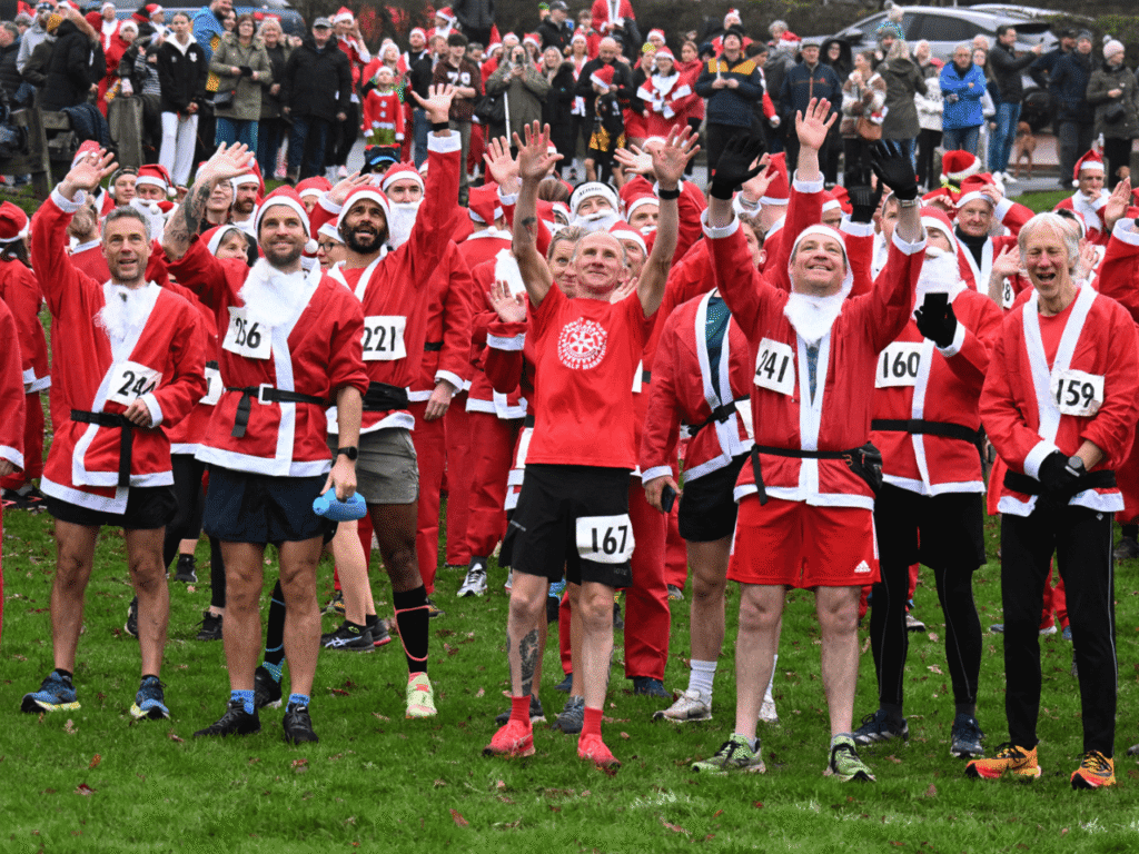 a group of people wearing santa suits stood with hands in the air on grass at the kemp santa fun run