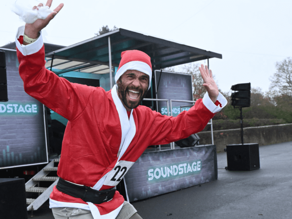 a man stand with his arms in the air wearing a santa suit top at the kemp santa fun run