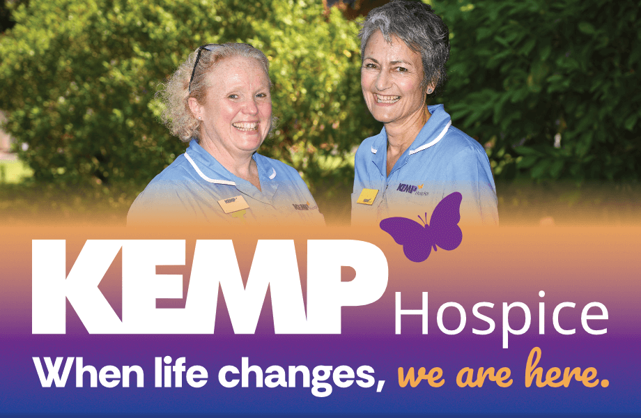 two ladies wearing light blue nurse scrubs smile at the camera whilst stood infront of bushes
