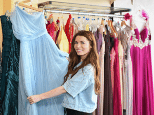 a lady with a denim jacket and brown hair smiles at the camera as she shows a light blue formal dress