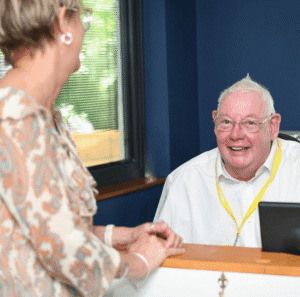 a gentleman with a white shirt wears a yellow lanyard and is smiling at a reception desk.
