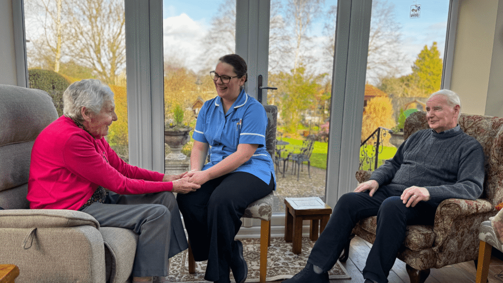 a lady sits in an armchair on the left wearing a bright pink cardigan whilst holding hands with a nurse in the centre of the screen who is wearing blue scrubs. A gentleman sits in an armchair to the right of the picture wearing a dark jumper.