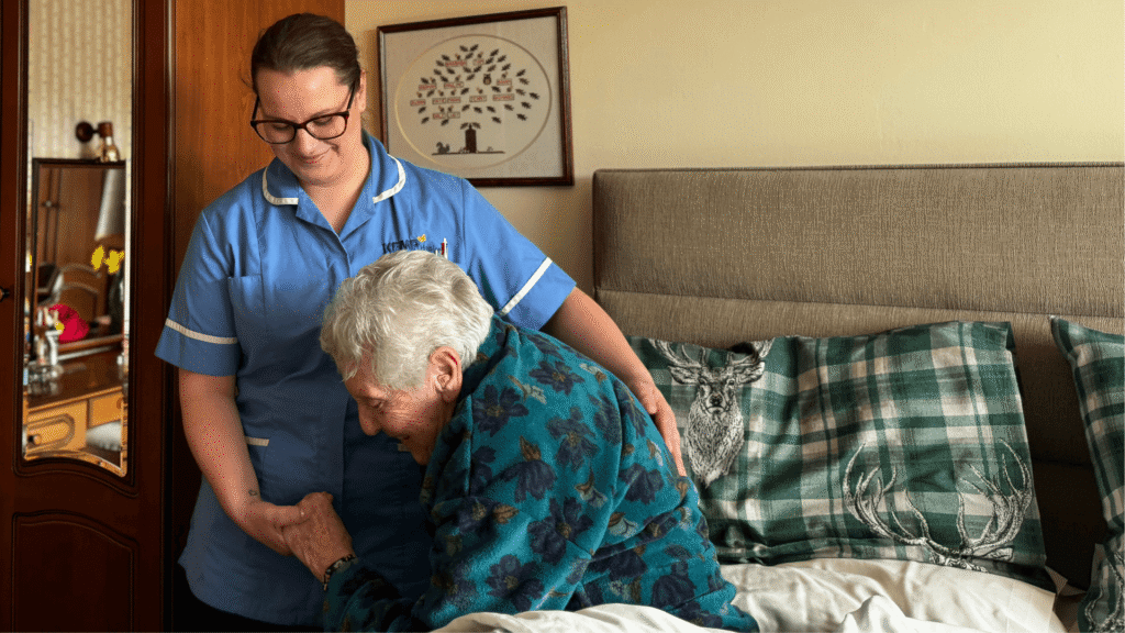 a nurse wearing blue scrubs helps another lady as she gets out of bed.