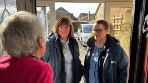 two nurses wearing kemp jackets stand at the door of a home being welcomed in by a lady wearing a pink cardigan.
