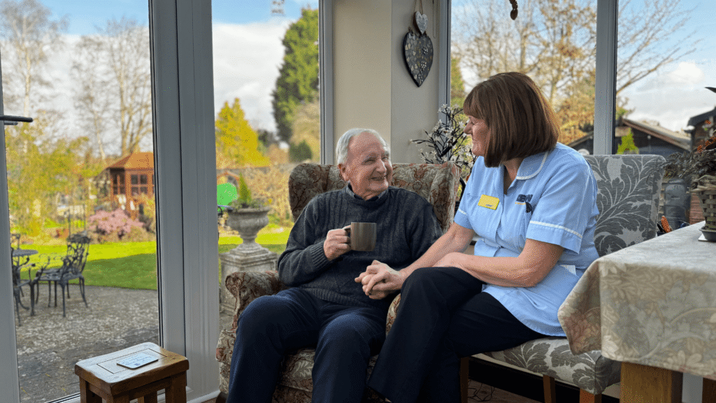 a gentleman sits in an armchair with a garden in the background. He holds a cup of tea whilst a nurse in blue scrubs holds his hand.