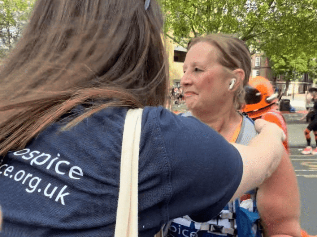 A lady wearing a running vest at london marathon gets a hug from another lady wearing a kemp hospice t shirt