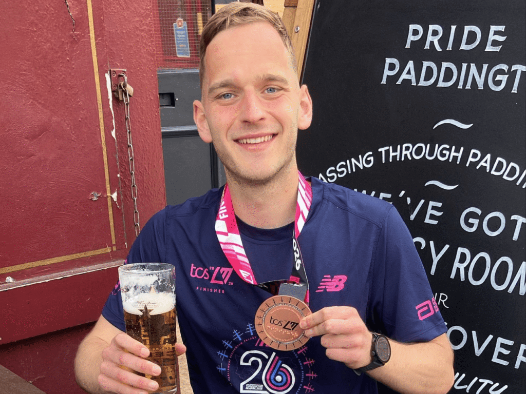 a man sits at a bench holding a pint of beer in one hand and a london marathon finisher medal in the other