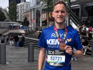 a gentleman wearing a kemp hospice t shirt with a London marathon finishers medal around his neck. Small crowds of people are in the background.