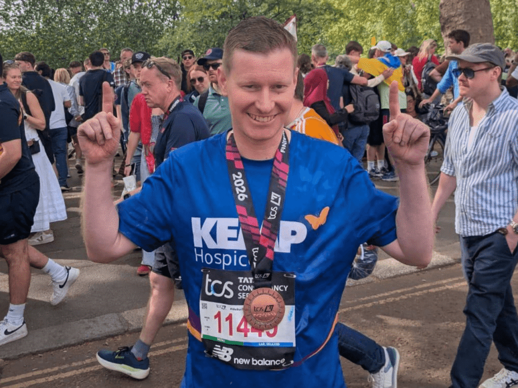 a gentleman wearing a kemp hospice t shirt holds his fingers in the air in a 'number one' gesture with a London marathon finishers medal around his neck. Crowds of people are in the background.