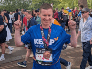 a gentleman wearing a kemp hospice t shirt holds his fingers in the air in a 'number one' gesture with a London marathon finishers medal around his neck. Crowds of people are in the background.