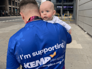 a man holds his baby on his shoulder at the london marathon whilst wearing a blue kemp t shirt. The picture is taken from the mans back with the baby facing the camera.