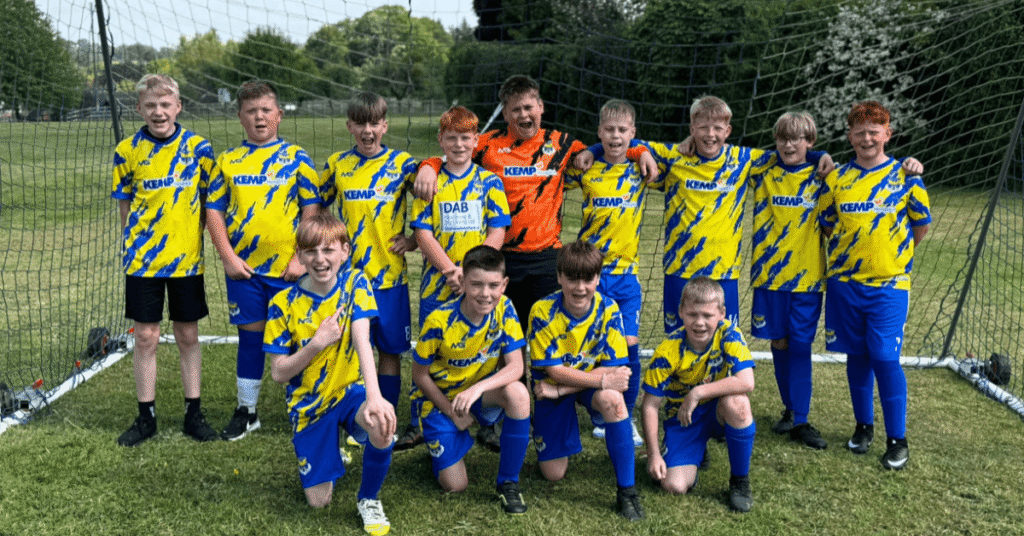 a groups of young people stand in a goal wearing football kits with KEMP Hospice sponsorship