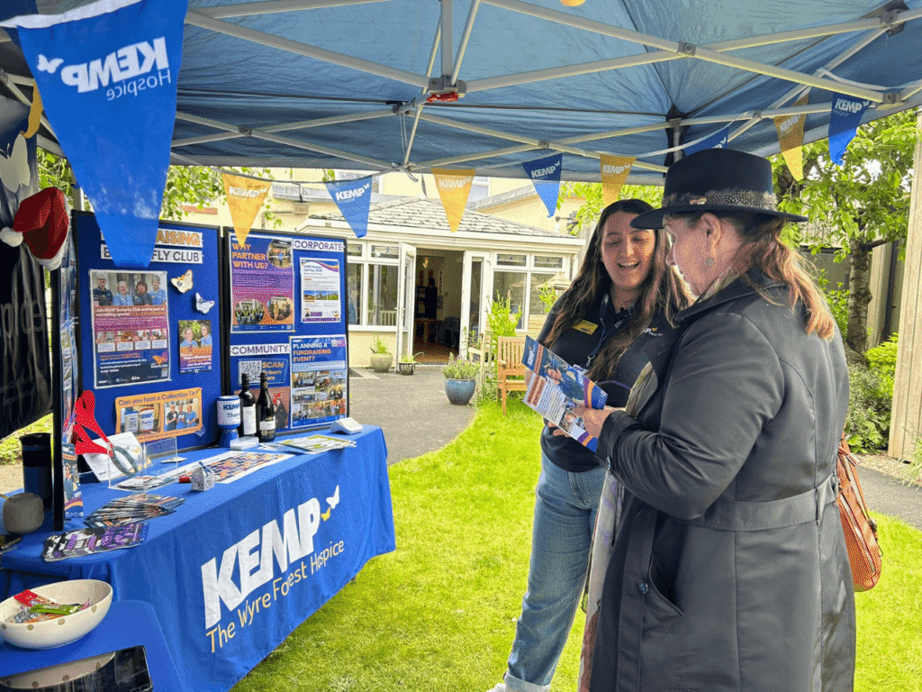 two ladies stand under a gazebo in a garden looking at a leaflet and talking