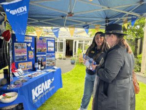 two ladies stand under a gazebo in a garden looking at a leaflet and talking