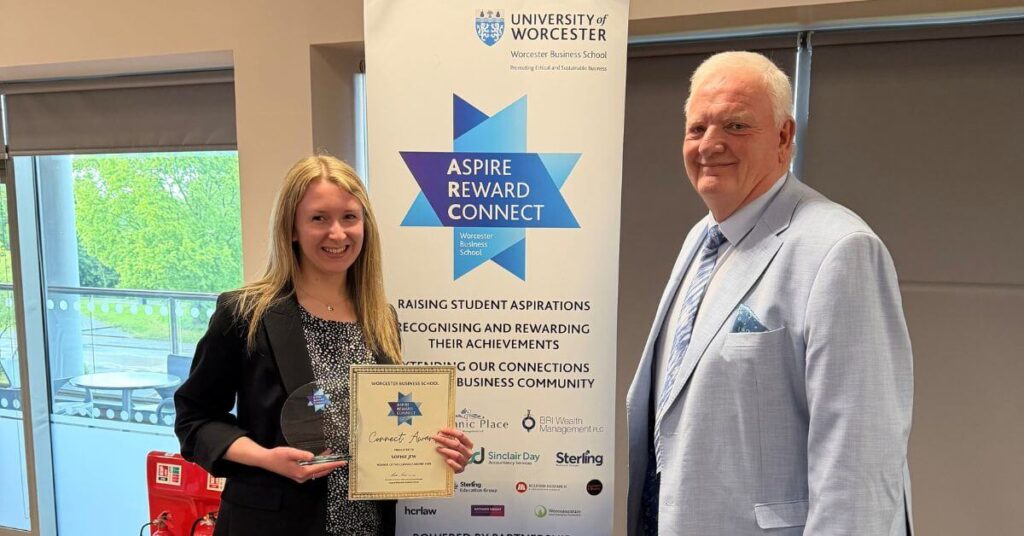 a lady stands holding a certificate and an award. a gentleman stands next to her on the right hand side. both are smiling.