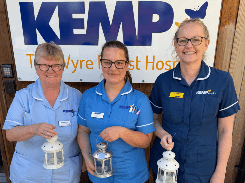 three ladies wearing nurse scrubs stand in front of a kemp hospice sign on a building. Each holds a lantern