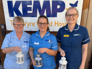 three ladies wearing nurse scrubs stand in front of a kemp hospice sign on a building. Each holds a lantern