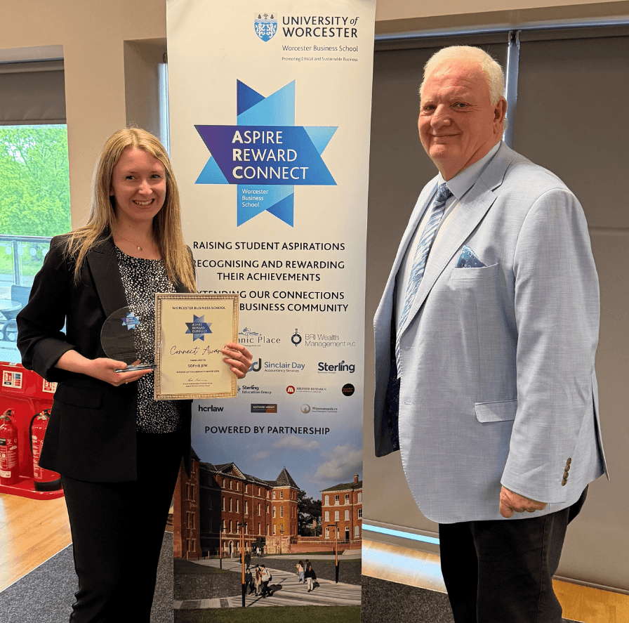 a lady stands holding a certificate and an award. a gentleman stands next to her on the right hand side. both are smiling.