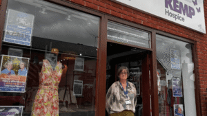 a lady in a green and white top stands in the open doorway of the boutique by kemp hospice
