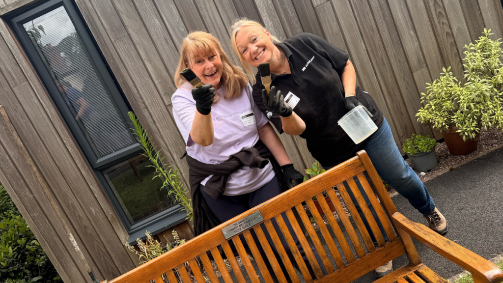 two people with paint brushes and a paint pot in their hands stood behind a wooden bench