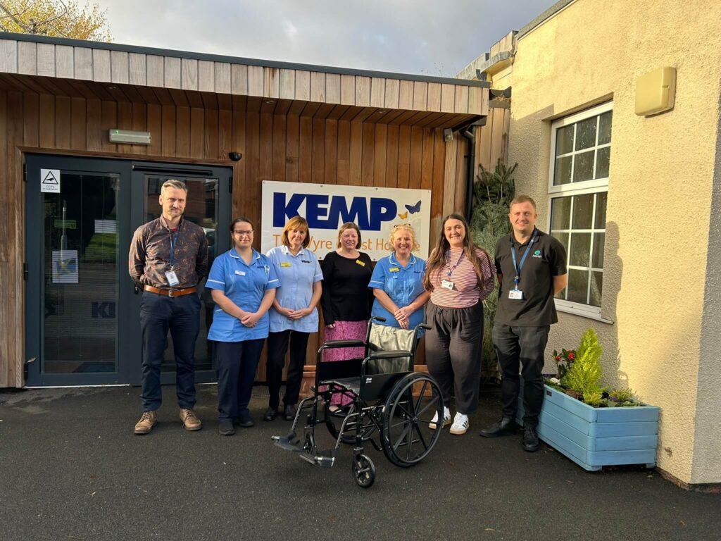 Group of nurses, corporate supporters and staff smiling in front of a hospice building with a wheelchair that was donated