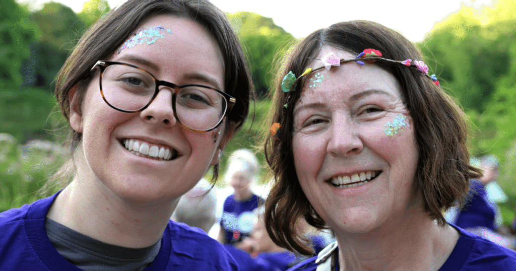 two peoples smiling at the camera with one wearing a flower garland headband and both with glitter paint on their faces.