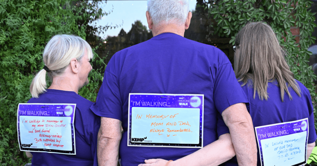 three people stand with their backs to the camera, arm in arm, with messages to loved ones on cards pinned to their backs