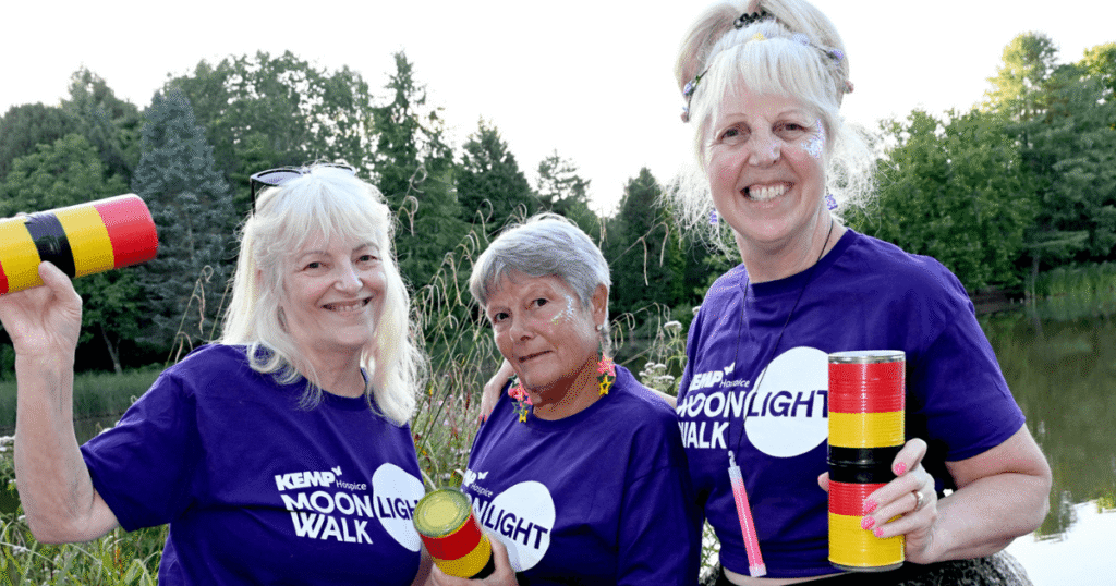 three people stand smiling at the camera with a lake in the background. They are wearing purple t shirts with kemp moonlight walk