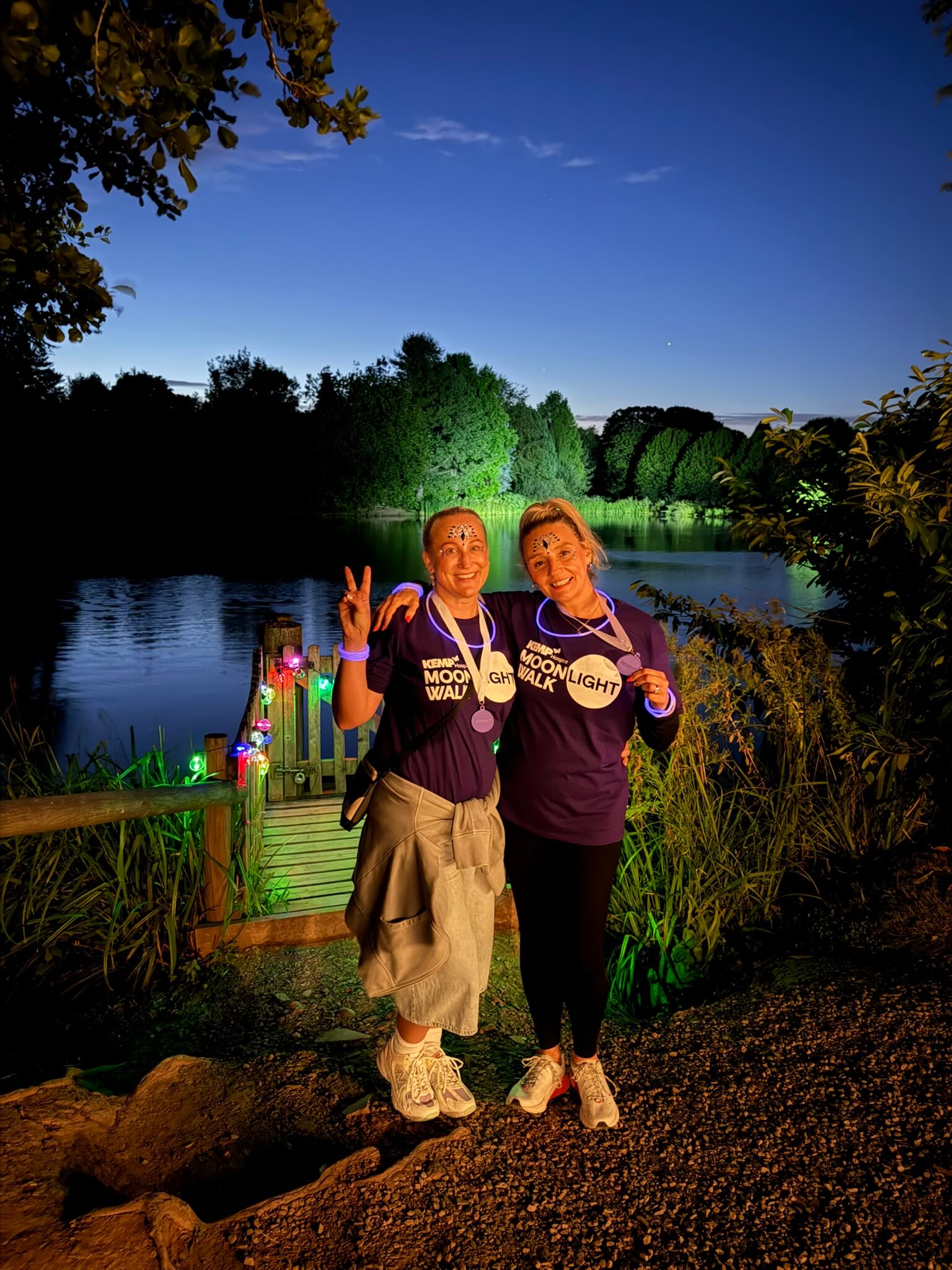 two people stand by a lake which is lit up at night. Both are wearing purple t shirts with kemp moonlight walk written on the front and both have purple medals.