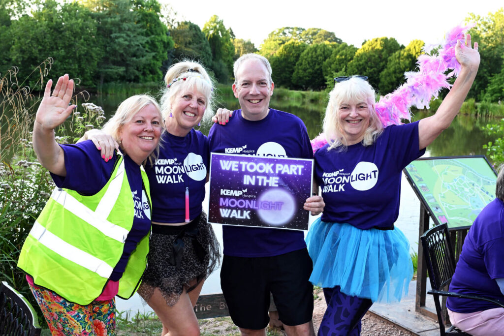 A group of four people stand together by a lake holding a sigh saying we took part in the kemp hospice Moonlight walk together