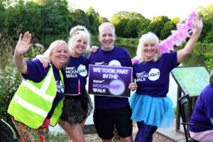 A group of four people stand together by a lake holding a sigh saying we took part in the kemp hospice Moonlight walk together