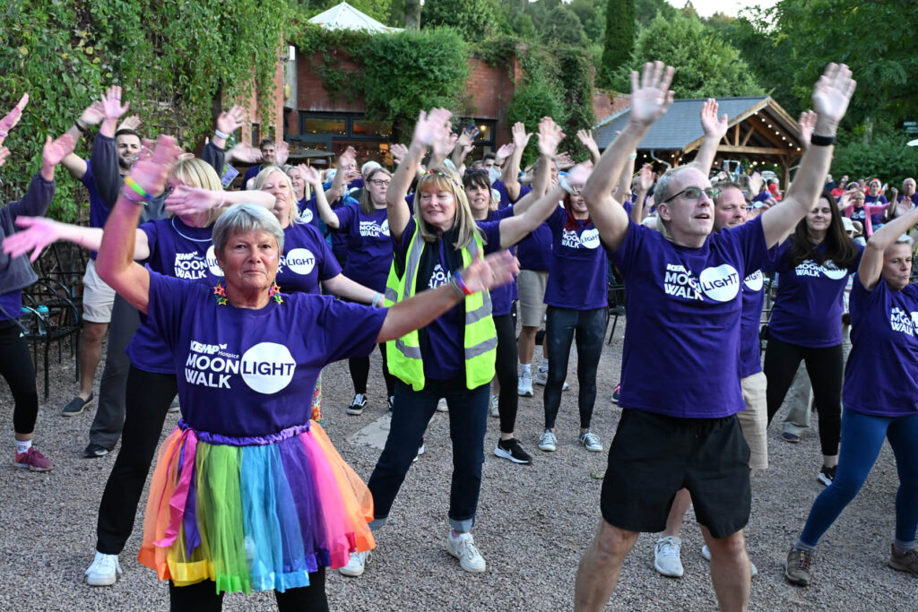 A group of people warming up for the kemp moonlight walk. They are wearing purple t shirts with kemp moonlight walk and some are wearing neon brightly coloured clothing
