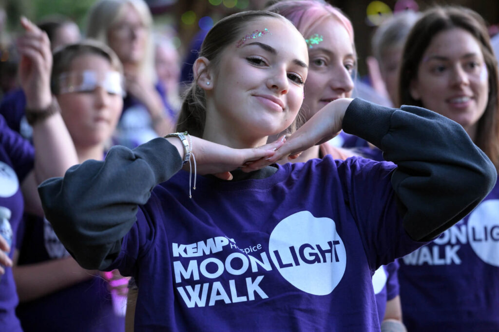 A person smiling at the camera with their hands placed under their chin to frame their face. They are wearing a purple t shirt with kemp moonlight walk on he front.
