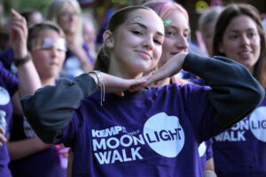 A person smiling at the camera with their hands placed under their chin to frame their face. They are wearing a purple t shirt with kemp moonlight walk on he front.