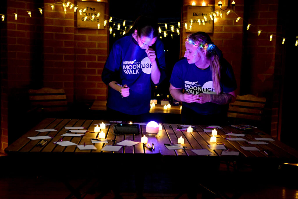 two people stand in a room which is gently lit by tea lights and battery candles. They are writing a message in a decorated card to be hung with other messages along the lights.